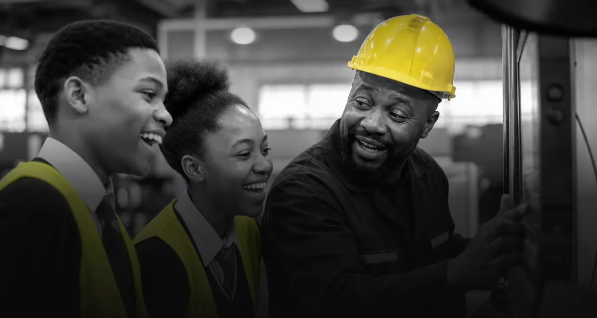 Two learners in hi-vis vests laughing as an engineer in a hard hat shows them a machine