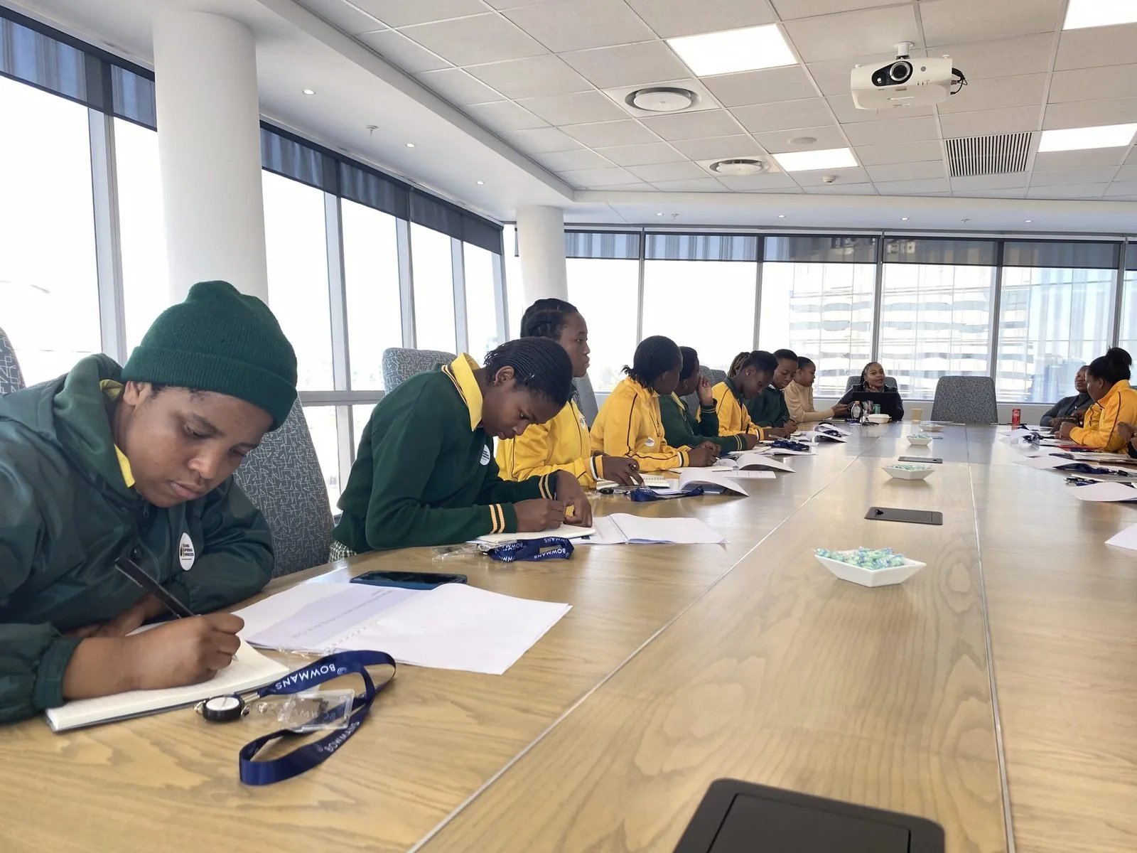 Learners in school tracksuits taking notes around a boardroom table on a workplace visit