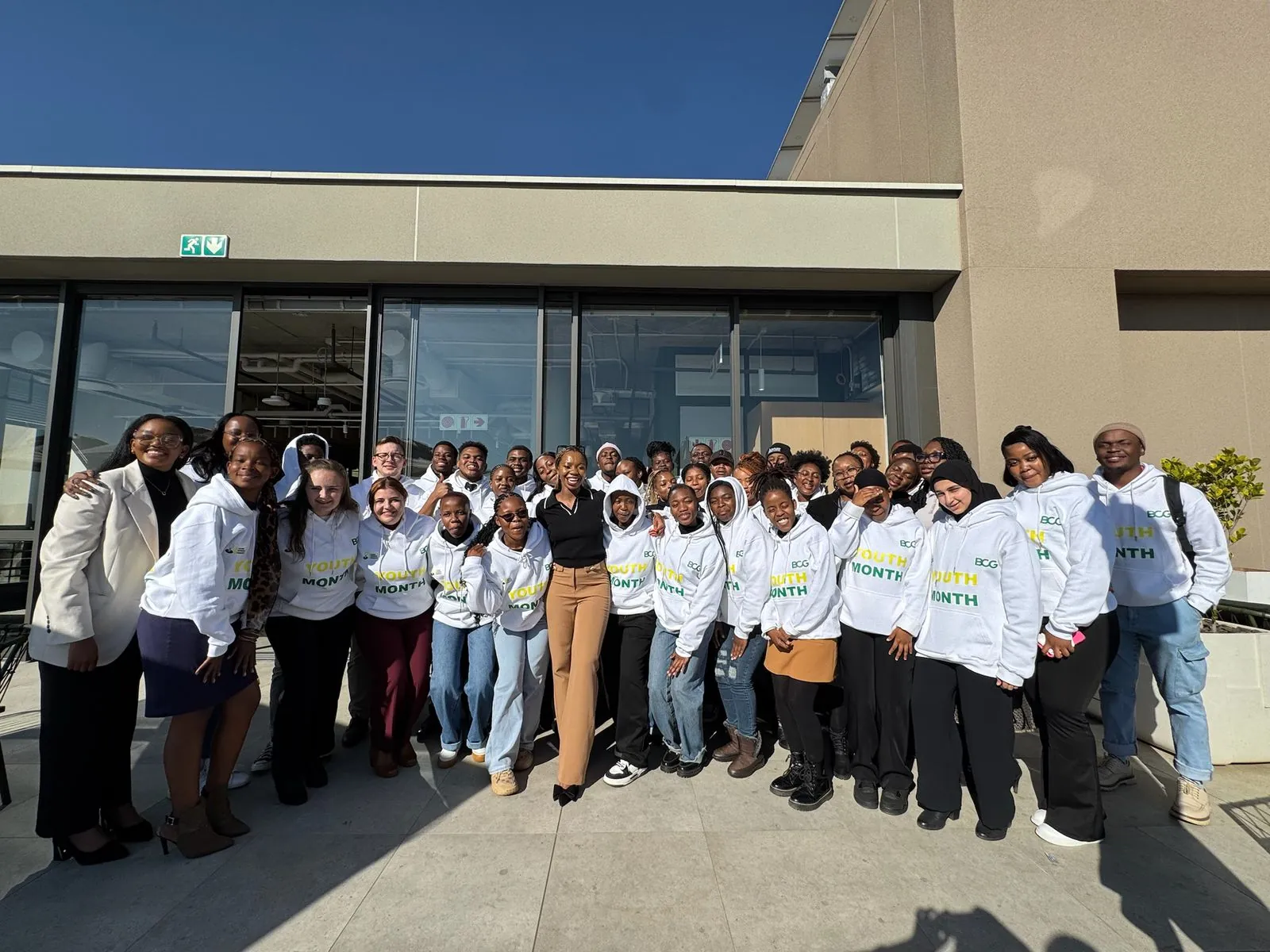 A large group of graduates in matching hoodies outside a campus building with their facilitators
